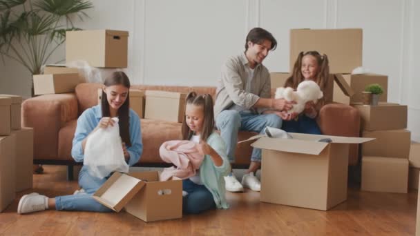 Grote verhuisdag. Gelukkig gezin van vier uitpakken dozen in nieuw huis, vrolijke kleine tweelingzusjes helpen ouders — Stockvideo