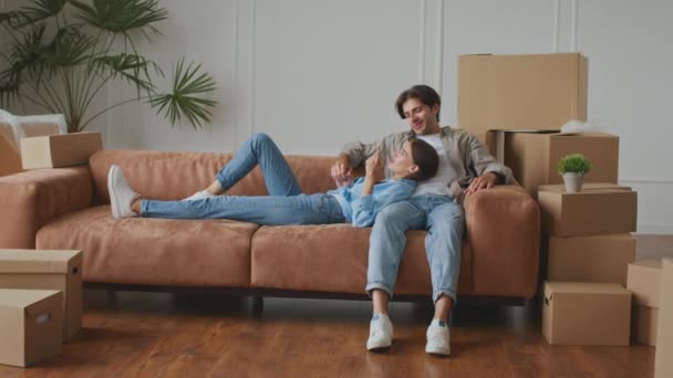 Nyt hjemmekoncept. Unge glade ægtefæller hviler på sofaen i ny lejlighed blandt bevægelige kasser og diskutere interiør – Stock-video