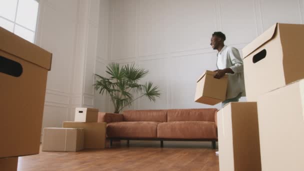 Koncept přemísťování. Šťastný mladý africký Američan nesoucí krabici s věcmi, tancující a sedící na pohovce — Stock video