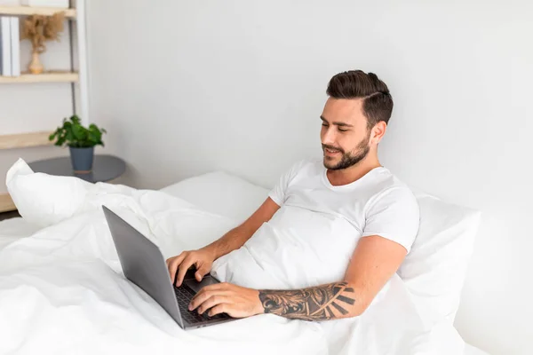 Praca w sypialni, surfing, przeglądanie w Internecie. Szczęśliwy człowiek tysiąclecia obudził się, siedząc na białym łóżku, wolna przestrzeń — Zdjęcie stockowe