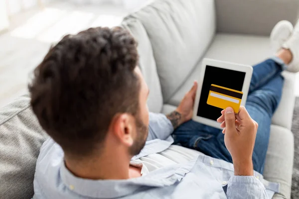 Έννοια του ηλεκτρονικού εμπορίου. Αγνώριστος άνθρωπος με πιστωτική κάρτα και ψηφιακό tablet κάθεται στον καναπέ στο σπίτι, πάνω από πυροβολισμό στον ώμο — Φωτογραφία Αρχείου