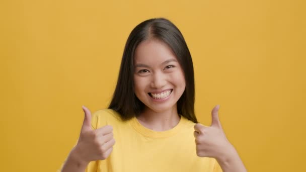 Sonriente señora asiática gestos Pulgares hacia arriba con ambas manos, fondo amarillo — Vídeo de stock