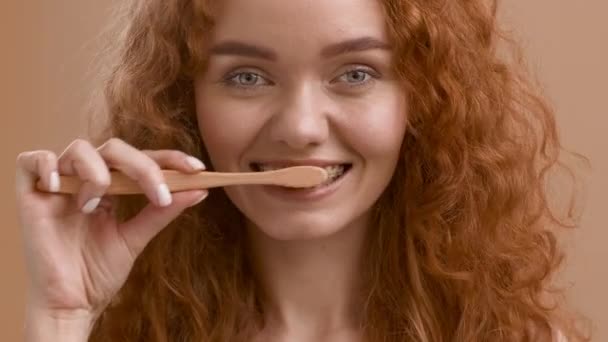 Mujer pelirroja con tirantes que limpian los dientes con cepillo de dientes, fondo beige — Vídeo de stock