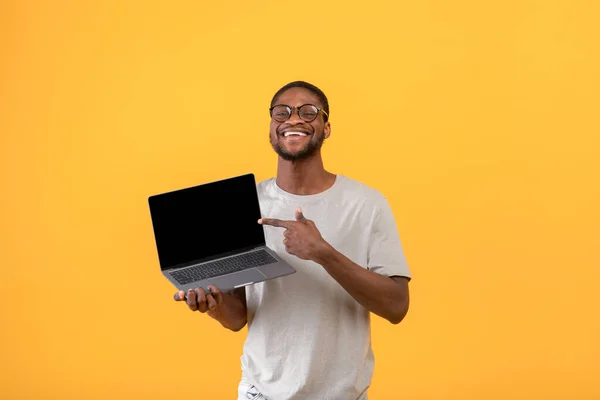 Hombre negro alegre apuntando a la pantalla del ordenador portátil en blanco con espacio para el diseño de su sitio web sobre fondo amarillo, maqueta — Foto de Stock