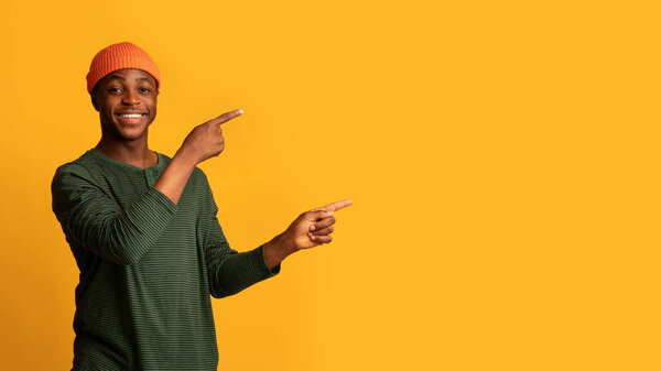 Check This. Smiling young black hispter guy pointing aside with two hands, cheerful african american man demonstrating copy space on yellow background, showing free place for advertisement, panorama
