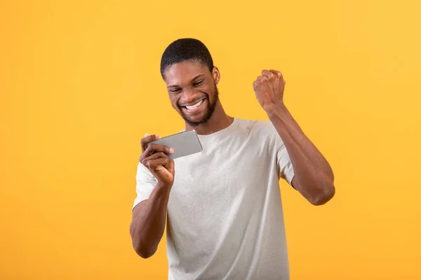Overjoyed uomo nero che celebra la vittoria con smartphone, ha vinto la lotteria online o gioco, sorridendo su sfondo giallo — Foto Stock