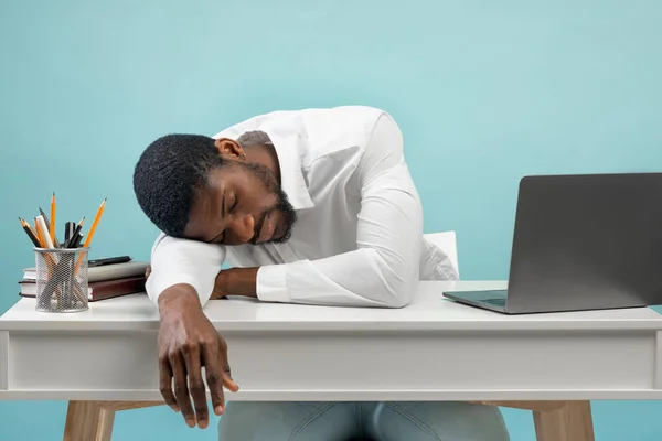 Jonge uitgeput zwarte werknemer slapen op de werkplek, moe van het werk en zet hoofd op tafel, blauwe achtergrond — Stockfoto
