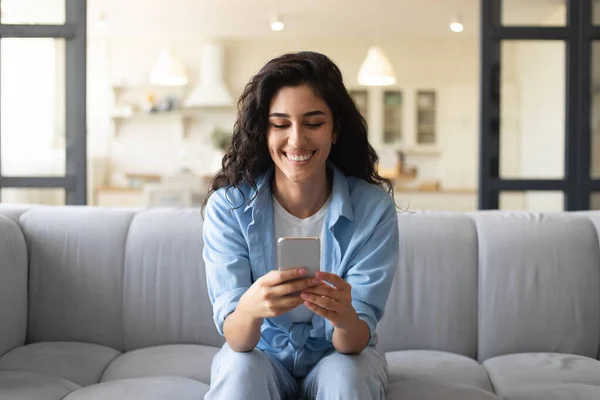 Giovane donna felice seduta sul divano con smartphone, controllare i social media, navigare in internet, chattare online da casa — Foto Stock