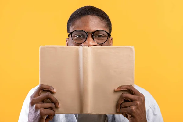 Eğitim konsepti. Gözlüklü Afro-Amerikalı adam yüzünü kitapla kapatıyor, sarı arka planda saklanıyor. — Stok fotoğraf