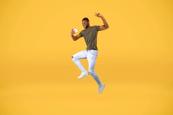 Çılgın Afrikalı Amerikalı adam, sarı stüdyo arka planında havada zıplarken akıllı telefondan selfie çekiyor. — Stok fotoğraf