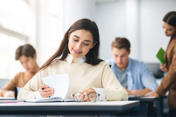 Estudante sorrindo sentado na mesa em sala de aula escrevendo em caderno — Fotografia de Stock