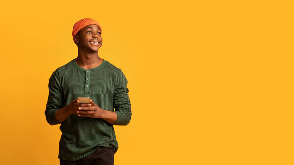 Dreamy Young Black Guy Holding Smartphone In Hands And Looking Aside At Copy Space, Pensive Millennial African American Man Using Cellphone While Standing Isolated Over Yellow Background, Panorama