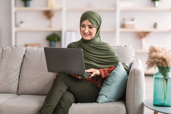 Веселая зрелая мусульманка с ноутбуком сидит на диване в помещении — стоковое фото