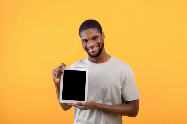 Młody czarny facet pokazuje cyfrowy tablet z czystym ekranem, polecając stronę internetową lub aplikację mobilną, żółte tło, — Zdjęcie stockowe