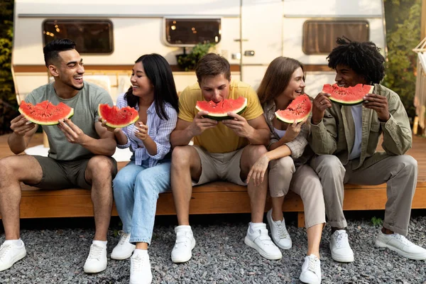 Alegre diversos amigos jóvenes comiendo sandía jugosa fresca, sentado cerca de autocaravana en viaje de otoño al campo — Foto de Stock