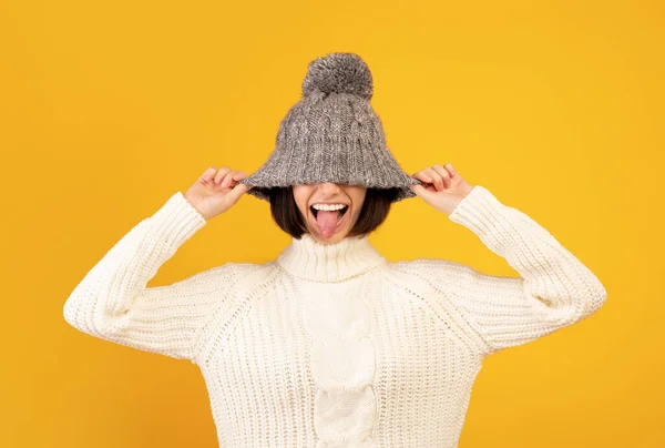 Hravá tisíciletá žena stahuje vlněný klobouk a vystrkuje jazyk, pohrává si se žlutým pozadím — Stock fotografie