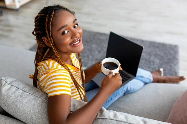 Señora negra positiva con taza de café usando el ordenador portátil, maqueta —  Fotos de Stock