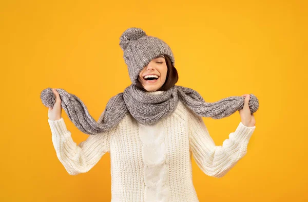 Jovem alegre com roupas quentes de inverno puxando para baixo chapéu de malha e brincando com cachecol no fundo amarelo — Fotografia de Stock