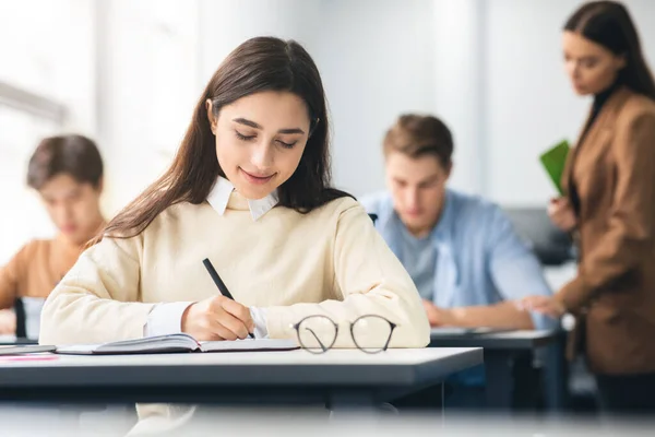 Mulher sorridente sentada à mesa em sala de aula escrevendo em caderno — Fotografia de Stock