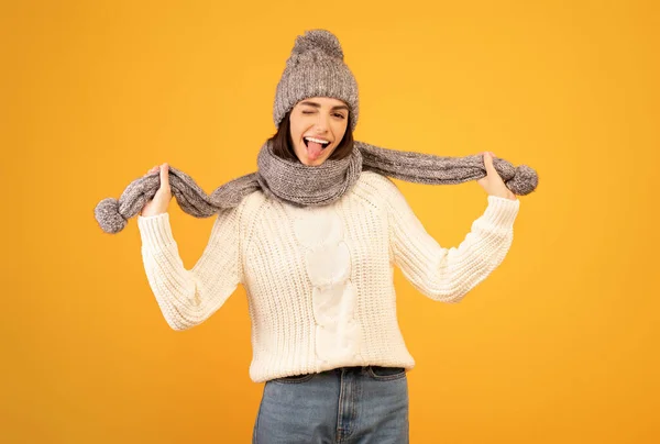 Senhora alegre em chapéu de gorro de malha cinza e cachecol saindo de sua língua e se divertindo no fundo do estúdio amarelo — Fotografia de Stock