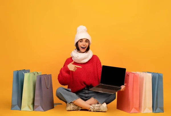 Mujer adicta a las compras feliz en traje de invierno apuntando a la pantalla del ordenador portátil, sentado entre bolsas de comprador, fondo amarillo — Foto de Stock