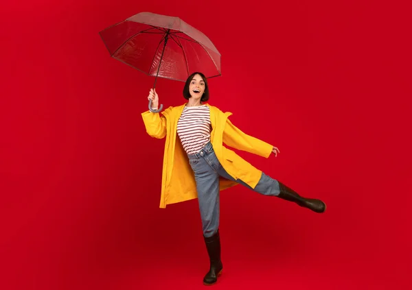 Playful young woman holding umbrella and having fun, laughing and looking at camera over red studio background — Stock Photo, Image