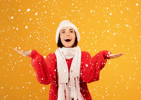 Kış geldi. Kış şapkalı ve eşarplı mutlu kadın kar taneleriyle oynuyor, sarı arka planda poz veriyor. — Stok fotoğraf