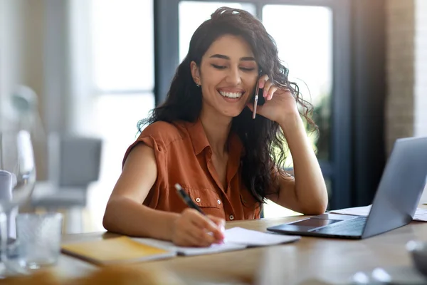 Concept de bureau à domicile. Joyeux jeune femme parlant sur smartphone, en utilisant un ordinateur portable, écrire des informations à l'intérieur — Photo