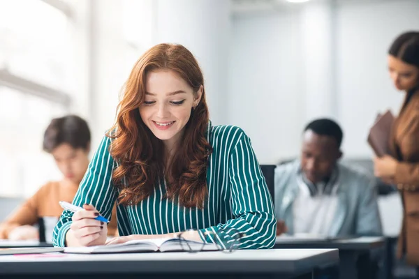 Dame souriante assise au bureau en classe écrivant dans un cahier — Photo