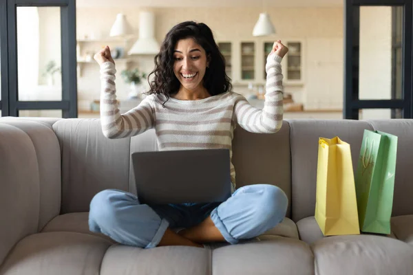 Mujer joven emocionada con el ordenador portátil y bolsas de compras haciendo el gesto SI, la compra de cosas en línea desde casa — Foto de Stock