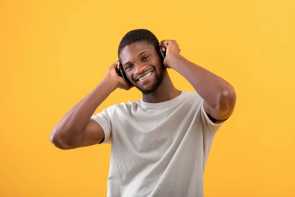 Ritratto di uomo afroamericano positivo che ascolta musica in cuffie senza fili, posa su sfondo giallo — Foto Stock