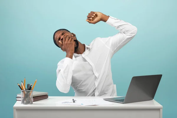 Hombre de negocios afroamericano bostezando y estirando las manos, mirando soñoliento, sentado en el lugar de trabajo, fondo azul —  Fotos de Stock