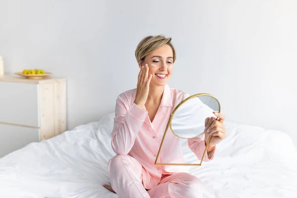 Portrait of smiling woman holding mirror applying cream on cheeks — Stock Photo, Image