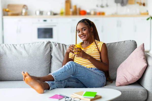 Mujer negra feliz sentada en el sofá en casa, bebiendo café —  Fotos de Stock