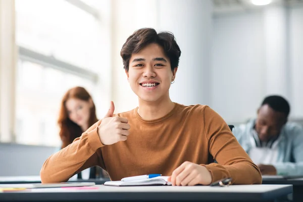 Asiático macho estudiante sentado en escritorio mostrando pulgares arriba gesto —  Fotos de Stock