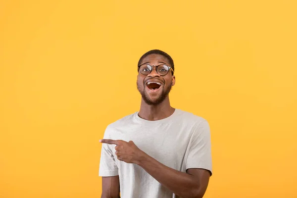 Sorprendido afroamericano hombre apuntando a un lado en el espacio en blanco y mirando a la cámara sobre fondo amarillo estudio —  Fotos de Stock