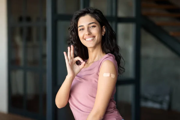 Junge Frau mit Pflasterhilfe am Arm nach Coronavirus-Impfung zeigt Okay und empfiehlt Covid-19-Impfung zu Hause — Stockfoto