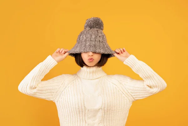 Mooie jonge vrouw trekken wollen hoed, tonen kussen lippen, gek en poseren voor camera over gele achtergrond — Stockfoto