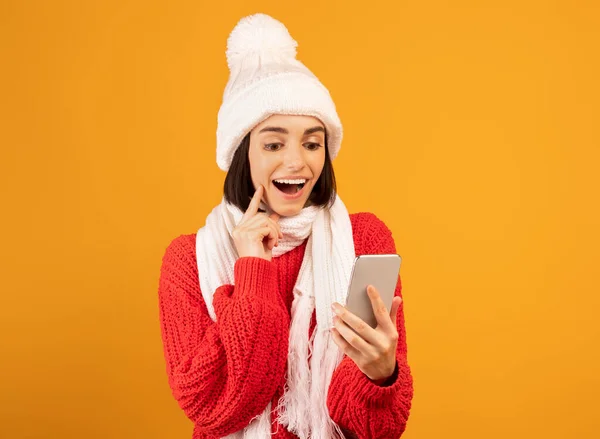 Concepto de compras de vacaciones de invierno remoto. Mujer excitada en sombrero de punto y bufanda mirando el teléfono inteligente, fondo amarillo — Foto de Stock