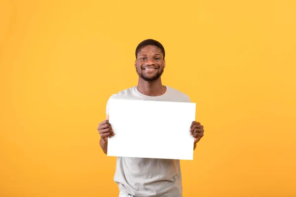 Fröhlicher afrikanisch-amerikanischer Mann mit weißem Blanko-Plakat, posiert und lächelt vor gelbem Studiohintergrund in die Kamera — Stockfoto