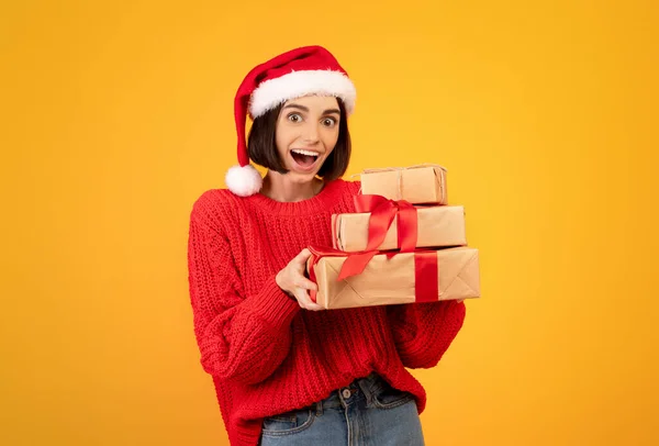 Eccitato signora in abito invernale e cappello di Babbo Natale contenente scatole regalo su sfondo giallo studio, spazio copia — Foto Stock