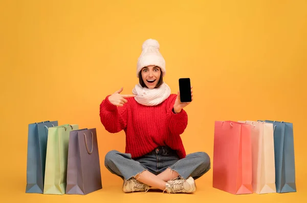 Aufgeregte Frau mit Wintermütze kauft online auf Smartphone ein und sitzt zwischen bunten Papiertüten auf gelbem Hintergrund — Stockfoto