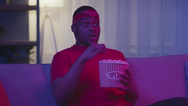 Африканский человек, смотрящий фильм ужасов, ест попкорн в помещении ночью — стоковое видео