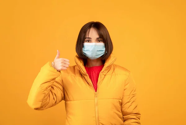 Счастливая юная леди в хирургической маске и зимней куртке показывает большой палец на желтом фоне — стоковое фото