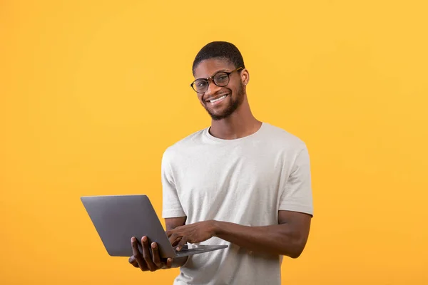 Ενθουσιασμένος μαύρος ελεύθερος επαγγελματίας που χρησιμοποιεί φορητό υπολογιστή, στέκεται στο στούντιο πάνω από κίτρινο φόντο και χαμογελώντας στην κάμερα, αντιγράφει χώρο — Φωτογραφία Αρχείου