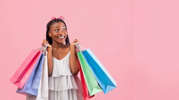 Alegre dama negra con bolsas de compras sonriendo y mirando el espacio vacío sobre fondo de estudio rosa, panorama —  Fotos de Stock