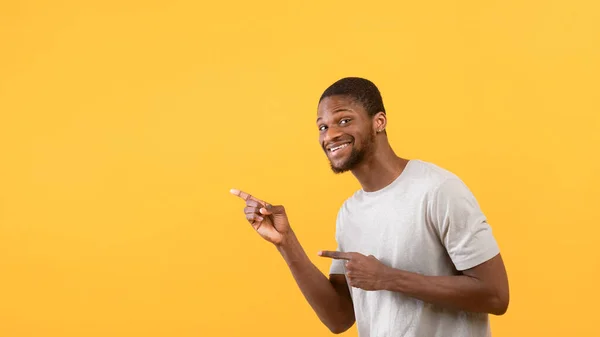 Emotionaler afrikanisch-amerikanischer Kerl zeigt mit dem Finger zur Seite auf leeren Raum, posiert und lächelt über gelbem Hintergrund — Stockfoto