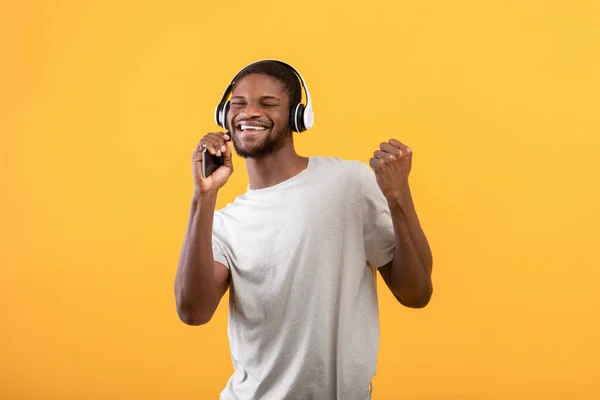 Tecnologías para la diversión. Chico afroamericano muy alegre escuchando música en auriculares y cantando sobre fondo amarillo — Foto de Stock