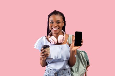 Sırt çantası, kulaklığı ve kahvesi olan mutlu siyah kız öğrenci pembe arka planda akıllı telefon gösteriyor.