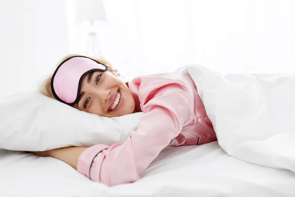 Счастливая кавказская женщина тысячелетия в розовой пижаме и маске для сна просыпается в уютной белой кровати с одеялом дома — стоковое фото